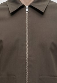 Gros plan d'une veste marron avec une fermeture éclair argentée à l'avant et un col pointu, portée par une personne avec un cou visible.