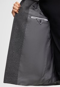 Innenseite eines grauen Wollmantels mit glattem grauen Futter, zwei Knöpfen und einer Tasche mit dreieckiger Klappe und Knopfakzent.