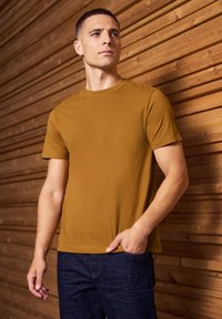 Hořčicově žluté bavlněné tričko s krátkým rukávem a kulatým výstřihem, spojené s tmavě modrými džíny proti dřevěnému pozadí.