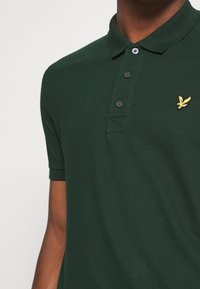 Dunkelgrünes Poloshirt mit einem Dreiknopfleisten, kurzen Ärmeln und einem gestickten gelben Vogel-Logo auf der linken Brust.