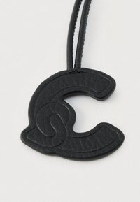 Μαύρο δερμάτινο μενταγιόν σε σχήμα στυλιζαρισμένου γράμματος C με ραφές, δεμένο σε μια λεπτή μαύρη κλωστή σε λευκό φόντο.