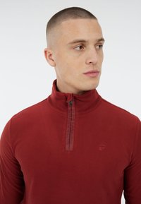 Pullover rosso in pile con colletto alto, chiusura a mezza zip con logo tono su tono e texture liscia.