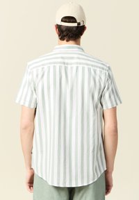 Camisa de manga corta en verde claro con rayas verticales en blanco, hecha de una tela suave. Presenta un dobladillo curvo y un detalle en el yugo del hombro.