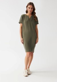Olive green short-sleeve dress with a V-neck, made from soft fabric. Features a relaxed fit and falls just above the knee. Paired with beige sneakers.