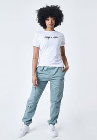 Witte t-shirt met zwart logo, gecombineerd met lichtblauwe cargobroek met zakken en elastische manchetten, aangevuld met witte sneakers.