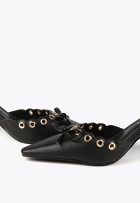 Svarta lädermules med spetsiga tår, scalloperade kanter, guldöglor och en rosettdetalj på vaden. Slät textur, slip-on design.