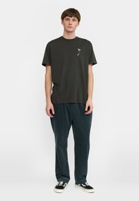 Camiseta de algodón verde oscuro con cuello redondo y mangas cortas, que presenta un pequeño gráfico floral en el lado izquierdo del pecho. Se lleva con pantalones sueltos y texturizados.