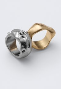 Δύο μεταλλικά δαχτυλίδια ακανόνιστου σχήματος, ένα ασημί και ένα χρυσό, τοποθετημένα δίπλα δίπλα σε απλό ανοιχτό φόντο.
