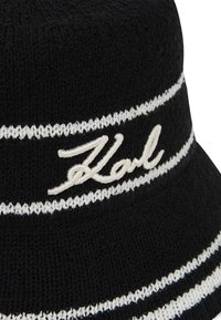 Cappello a secchiello nero lavorato a maglia con righe bianche, caratterizzato da un logo "Karl" ricamato. Tessuto morbido, forma rotonda, progettato per un uso casual.