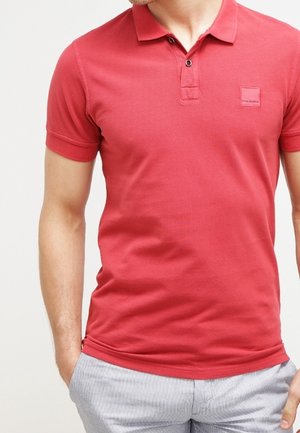 Mężczyzna ubrany w dopasowaną czerwoną koszulkę polo z dwoma guzikami i jasnoszare spodnie w paski, ręce w kieszeniach, na jednolitym tle.