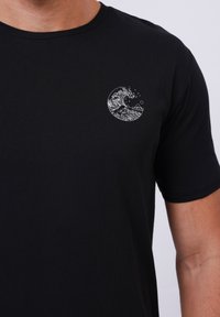 Μαύρο βαμβακερό T-shirt με κοντά μανίκια και στρογγυλή λαιμόκοψη. Διαθέτει ένα λευκό γραφικό με κυματισμούς στην αριστερή περιοχή του στήθους.