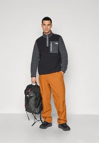 Črno-siv flis pulover z zadrgo na vratu, sivo prsno žepom, kombiniran z oranžnimi hlačami in črnim nahrbtnikom. Teksturirana tkanina in robustna zasnova.