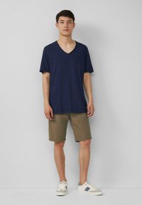 Granatowa koszulka polo z dekoltem w serek w lekkim materiale, zestawiona z oliwkowymi szortami. Model nosi białe sneakersy z szarymi akcentami. Prosty design.