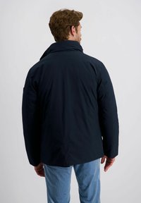 Mann mit kurzen braunen Haaren, der eine dunkelblaue Jacke und hellblaue Hose trägt, mit dem Rücken zu einem schlichten hellgrauen Hintergrund.