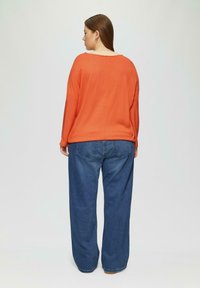 Oranje gebreide trui met een brede halslijn, gedragen met losse blauwe jeans met een klassiek vijf-pocket ontwerp en zichtbare stiksels.