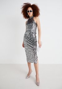 Srebrna obleka s sequins, ki ima visok ovratnik in prilegajočo silhueto, z naborki na strani in razporkom. V kombinaciji s prosojnimi petkami in clutch torbico.