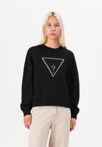 Crna dukserica od pamuka s spuštenim ramenima, koja u središtu ima trokutasti logo ukrašen rhinestones kamenčićima. Uparen s svijetlosmešavim hlačama.