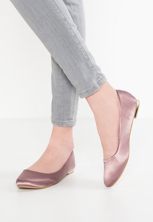 Fødder iført skinnende mauve balletflade sko kombineret med lysegrå skinny jeans med opsmøg på en hvid baggrund.