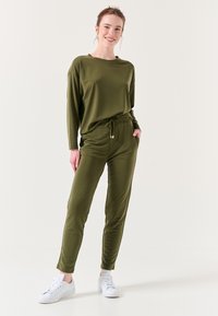 Olijfgroen loungewear set bestaande uit een losse langslijve top en taps toelopende broeken met een elastische tailleband en zakken, gecombineerd met witte sneakers.