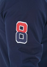 Chemise à manches longues bleu marine avec le numéro 8 brodé en rouge et blanc sur la manche gauche, présentant une texture en coton lisse.