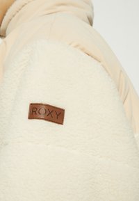 Roxy jacka i krämfärgad fleece, med släta ärmar i beige tyg och en brun läderlogotyp påsydd.