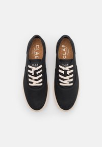 Černé plátěné tenisky s bílými tkaničkami, světle béžovými podrážkami a korekovými stélkami označenými "CLAE Los Angeles Vegan," pohled shora na bílém pozadí.