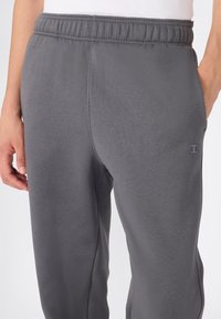 Close-up van een persoon in grijze sweatpants met elastische tailleband en het Champion-logo nabij de zak, met handen langs de zijden.