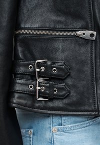 Κοντινό πλάνο στο πλαϊνό μέρος μαύρου δερμάτινου τζάκετ με ασημένιο φερμουάρ και δύο λουριά με αγκράφες, σε συνδυασμό με ανοιχτό μπλε τζιν παντελόνι.