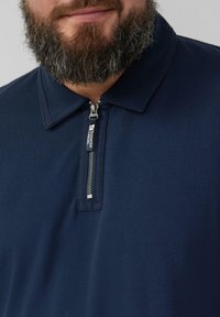 Granatowa koszulka polo z teksturowanej tkaniny, z zamkiem błyskawicznym przy kołnierzy i srebrnym detalem zamka. Brak widocznych wzorów lub akcentów.