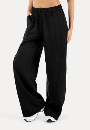 Person iført løse sorte joggingbukser med elastisk taljebånd og hvide sneakers, stående med den ene hånd i lommen.