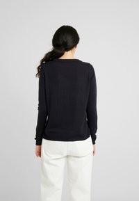 Maglione nero a maglia con maniche lunghe e scollatura rotonda, caratterizzato da polsini e orlo a coste, indossato sopra pantaloni di colore chiaro.