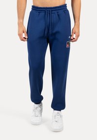 Navyblaue Baumwoll-Jogginghosen mit einem elastischen Bund und Kordelzug. Mit seitlichen Taschen und einem Logopatch am Oberschenkel. Kombiniert mit weißen Sneakern.