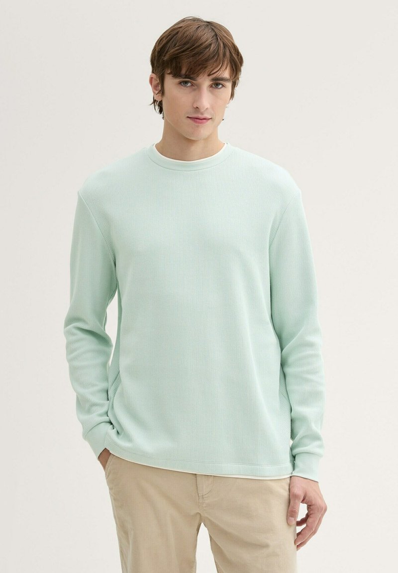 Helles mintgrünes langärmeliges Sweatshirt aus Rippstoff mit Rundhalsausschnitt, strukturiertem Stoff und lockerer Passform, kombiniert mit hellbeigen Hosen.