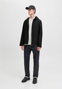 Młody mężczyzna w czarnej czapce, czarnej kurtce z kapturem, białej koszulce polo, ciemnych jeansach z podwiniętymi nogawkami i czarnych butach, stojący na jednolitym tle.