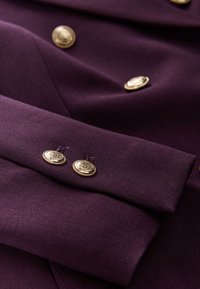 Lila Stoff mit glatter Textur, versehen mit goldfarbenen Knöpfen mit kunstvollen Verzierungen. Nahaufnahme eines gefalteten Abschnitts und der Knopfplatzierung.