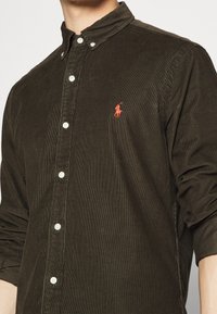 Mann in dunkelolivgrünem Cordhemd mit Knöpfen und hochgeschobenen Ärmeln, mit besticktem rotem Polo-Spieler-Logo auf der Brust.