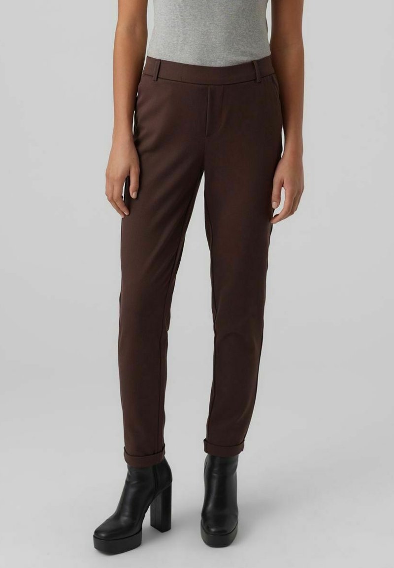 Femme portant un pantalon marron cintré avec des revers, des bottines noires à talons hauts, associées à un haut gris clair sans manches.