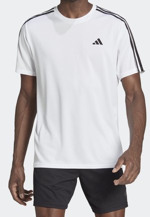 Mann trägt weißes Adidas Kurzarmshirt mit schwarzen Streifen auf den Schultern und schwarze Shorts, steht vor schlichtem Hintergrund.