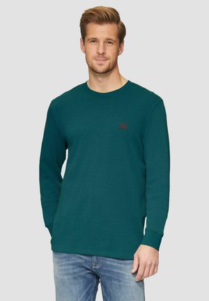 Chemise à manches longues teal avec un col rond et un petit logo noir sur la poitrine. Tissu doux avec une coupe classique, associée à un jean bleu clair.