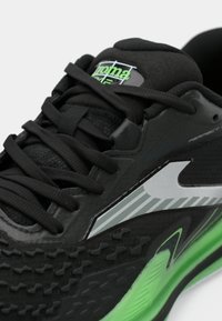 Čierna atletická obuv s textúrovaným horným dielom, zelenými akcentmi, reflexným strieborným pruhom a čiernymi šnúrkami. Obsahuje polstrovanú podrážku pre podporu.