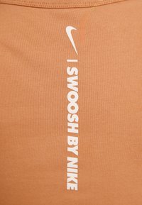 Pionowy biały napis "SWOOSH BY NIKE" oraz logo znaku Swoosh Nike na jasnobrązowym tle z tkaniny.