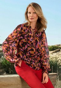 Camicia multicolore con un motivo floreale in rosa, arancione e nero, con maniche lunghe trasparenti e scollo rotondo, abbinata a pantaloni rossi.