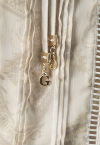 Tissu beige clair avec des plis complexes et deux fermoirs dorés, l'un orné d'une petite pierre précieuse et d'un pendentif en forme de lettre "G".