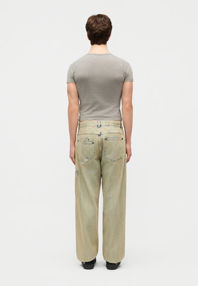 Άνδρας που στέκεται με την πλάτη, φορώντας εφαρμοστό γκρι μπλουζάκι, φαρδιά ανοιχτό μπεζ τζιν με τσέπες και μαύρα παπούτσια, σε λευκό απλό φόντο.