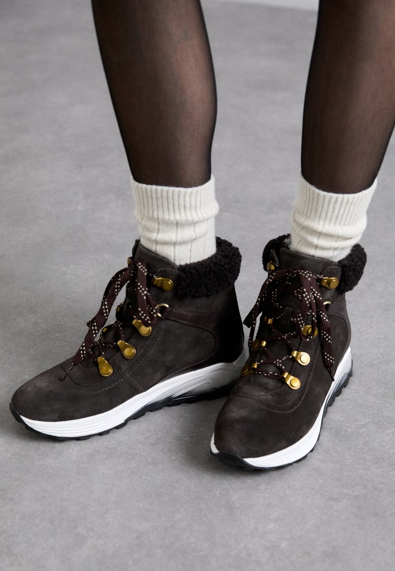 Botas de tobillo de ante marrón con suela blanca, puño forrado de piel, y cordones oscuros en contraste. Presentan herrajes dorados y un upper texturizado.