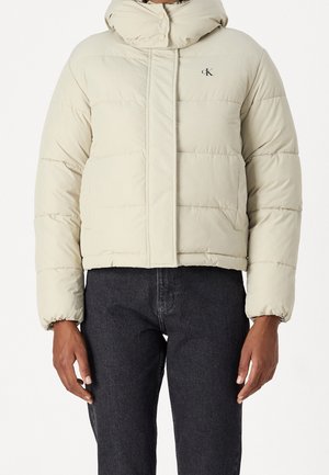 Doudoune courte beige avec un col montant et le logo Calvin Klein sur la poitrine, portée avec un jean noir.