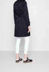 Marinblå trenchcoat med bälte i midjan, ryggslits och knappade ärmslut, kombinerad med vita ankellånga byxor och svarta sneakers med vita ränder.