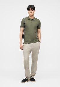 Άνδρας που φοράει πουκάμισο πόλο ελιάς, μπεζ παντελόνι με ρίγες, μαύρα παπούτσια χωρίς κορδόνια και μαύρη τσάντα ώμου, στέκεται με το ένα χέρι στην τσέπη.