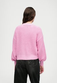 Lys rosa strikket genser med rund hals, dropped shoulders og ribbede mansjetter, paret med mørke, løstsittende bukser.