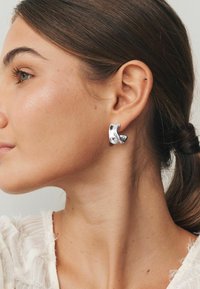 Sudraba auskari ar skulpturālu, viļņotu dizainu. Gluda tekstūra, spīdīgs pārklājums. Modelis tos valkā kreisajā ausī pret neitrālu fonu.